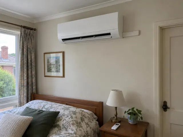 Bedroom wall with a high-mounted split system above a bed, curtains, lamp and side table in Mitcham